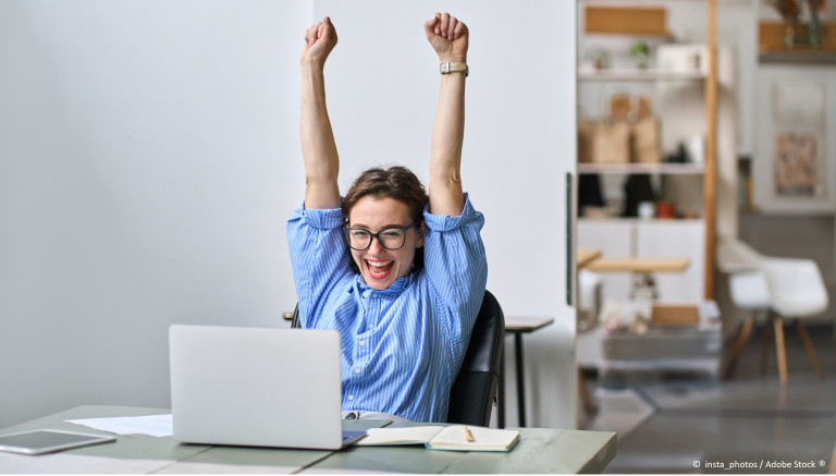 Une femme lève les bras en souriant suite à un succès sur ordinateur
